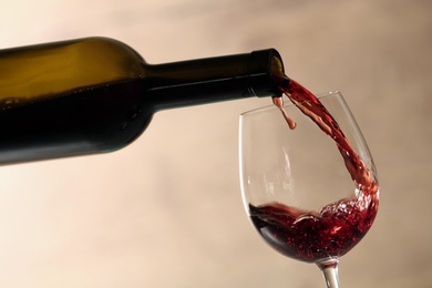 Pouring red wine into glass from bottle against blurred beige background, closeup