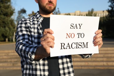 Photo of Young man holding sign with phrase Say No To Racism outdoors, closeup