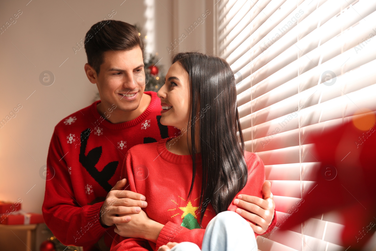 Photo of Happy couple in warm Christmas sweaters near window indoors