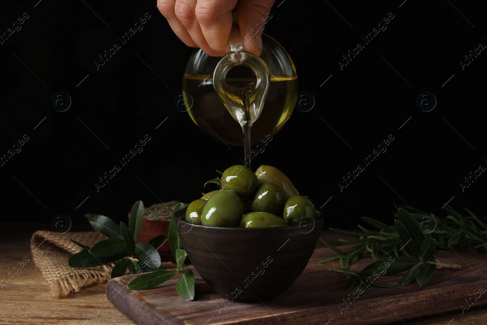 Photo of Woman pouring oil into bowl with ripe olives on wooden table, closeup