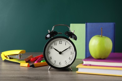 Photo of Alarm clock and different stationery on wooden table near green chalkboard. School time