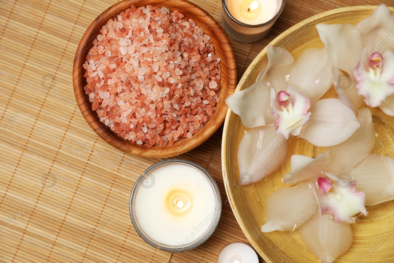 Photo of Bowl with water, flowers, sea salt and burning candles on bamboo mat, flat lay. Spa treatment