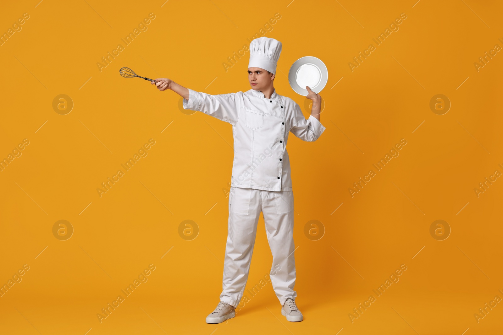 Photo of Portrait of confectioner in uniform holding plate and whisk on orange background