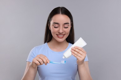 Photo of Happy woman squeezing toothpaste from tube onto plastic toothbrush on light grey background