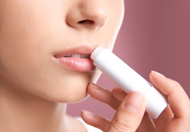 Young woman applying balm on her lips against color background, closeup