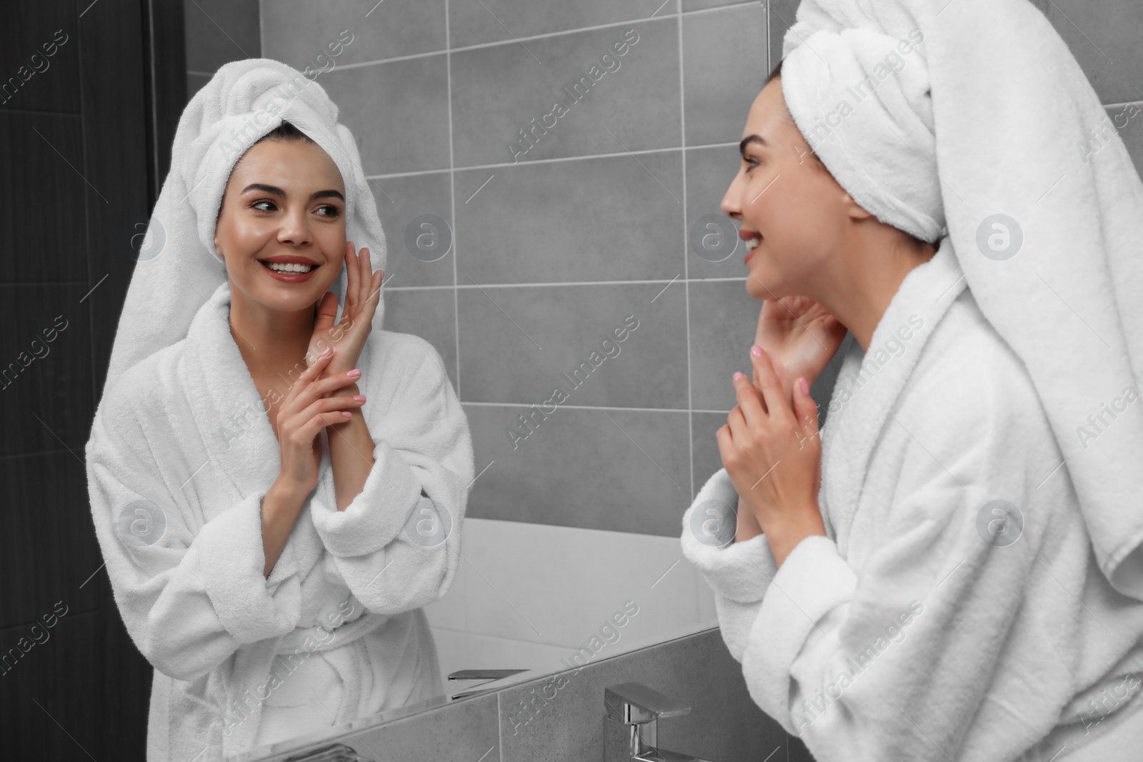 Photo of Beautiful young woman in bathrobe with towel on head near mirror at home
