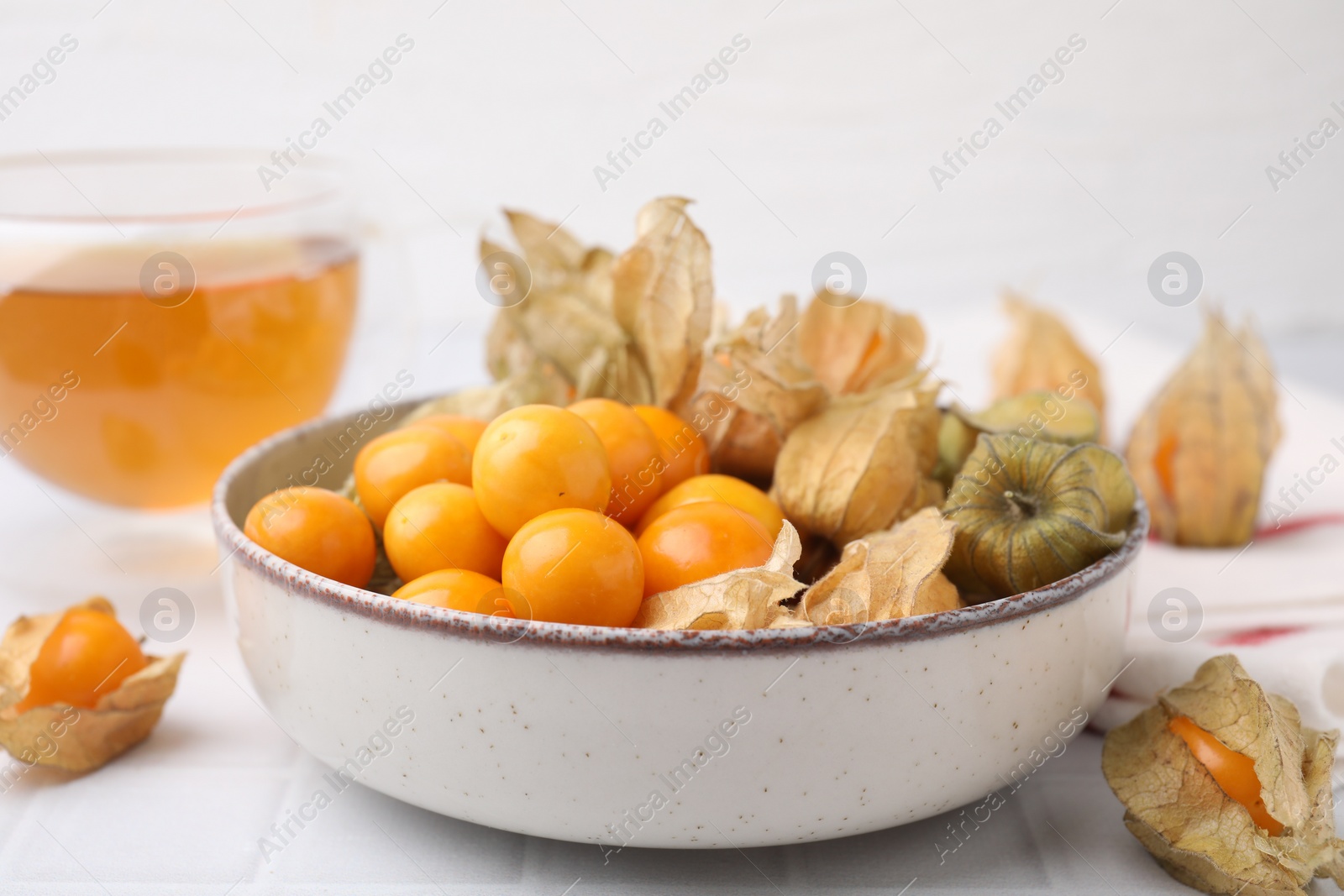 Photo of Ripe physalis fruits with calyxes in bowl on white tiled table, closeup