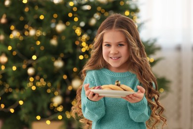Photo of Happy little girl holding plate with cookies near Christmas tree at home. Space for text