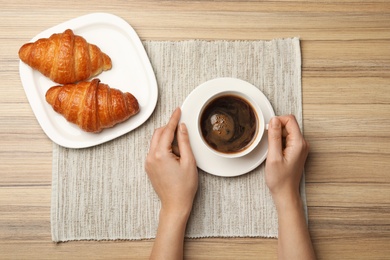 Woman with cup of coffee and croissants at wooden table, flat lay