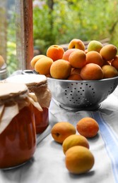 Photo of Delicious ripe apricots with jars of homemade jam on table near window