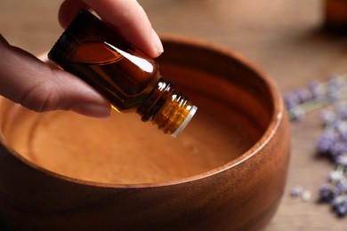 Woman dripping lavender essential oil from bottle into wooden bowl at table, closeup