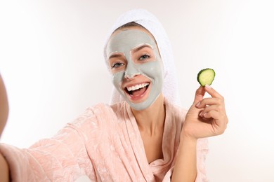 Photo of Woman with face mask and cucumber slice taking selfie on white background. Spa treatments