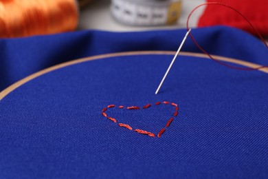 Photo of Red embroidered heart, thread and sewing needle on blue cloth with hoop, closeup
