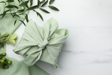 Photo of Furoshiki technique. Gift packed in green fabric and plants for decoration on white marble table, flat lay. Space for text