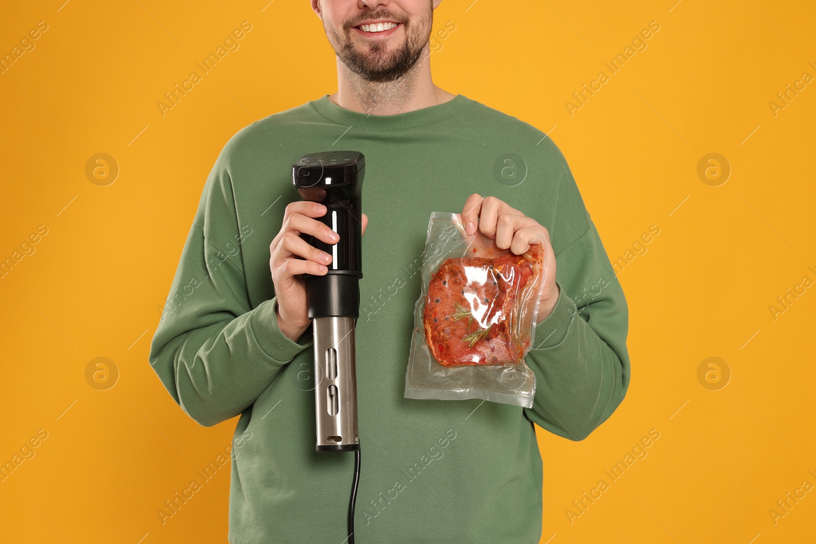 Photo of Smiling man holding sous vide cooker and meat in vacuum pack on orange background, closeup