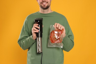 Photo of Smiling man holding sous vide cooker and meat in vacuum pack on orange background, closeup