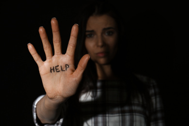 Abused young woman showing palm with word HELP against black background, focus on hand. Domestic violence concept