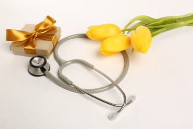 Photo of Stethoscope, gift box and yellow tulips on white background. Happy Doctor's Day