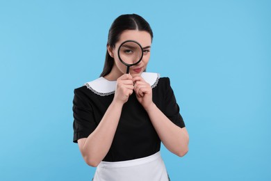 Photo of Young woman in maid outfit looking through magnifier glass on light blue background
