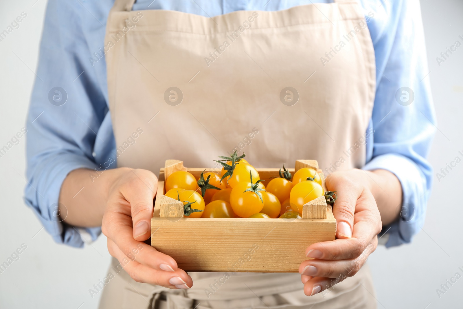 Photo of Woman holding wooden crate of yellow tomatoes on light background, closeup