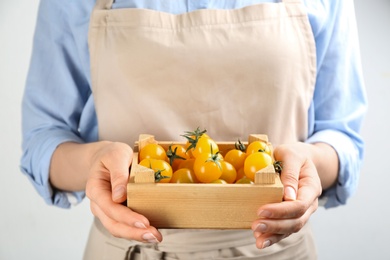 Photo of Woman holding wooden crate of yellow tomatoes on light background, closeup