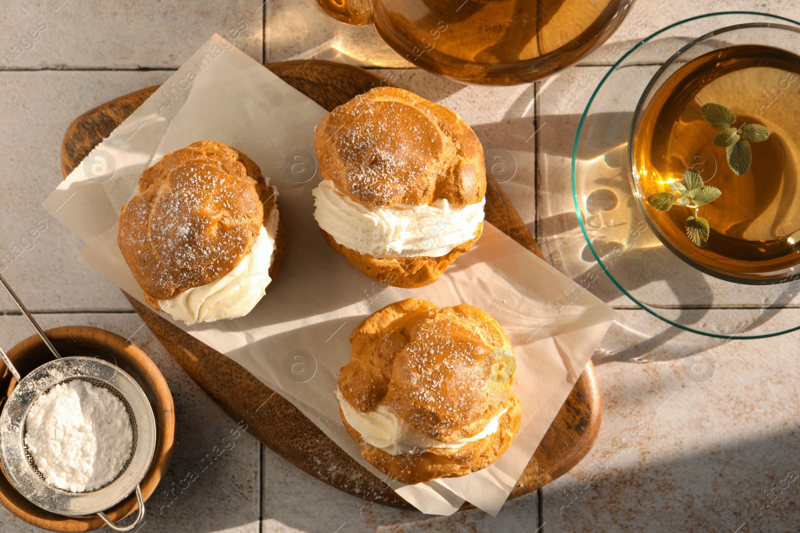 Photo of Delicious profiteroles filled with cream and tea on white tiled table, flat lay
