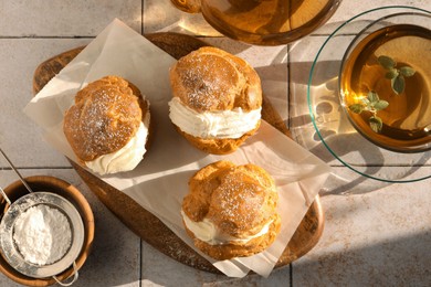 Delicious profiteroles filled with cream and tea on white tiled table, flat lay
