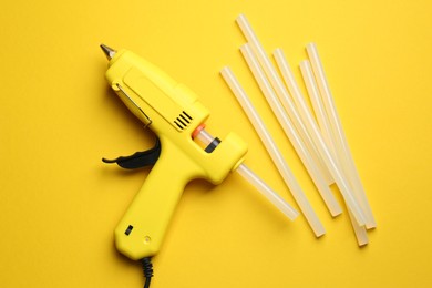 Glue gun and sticks on yellow background, flat lay