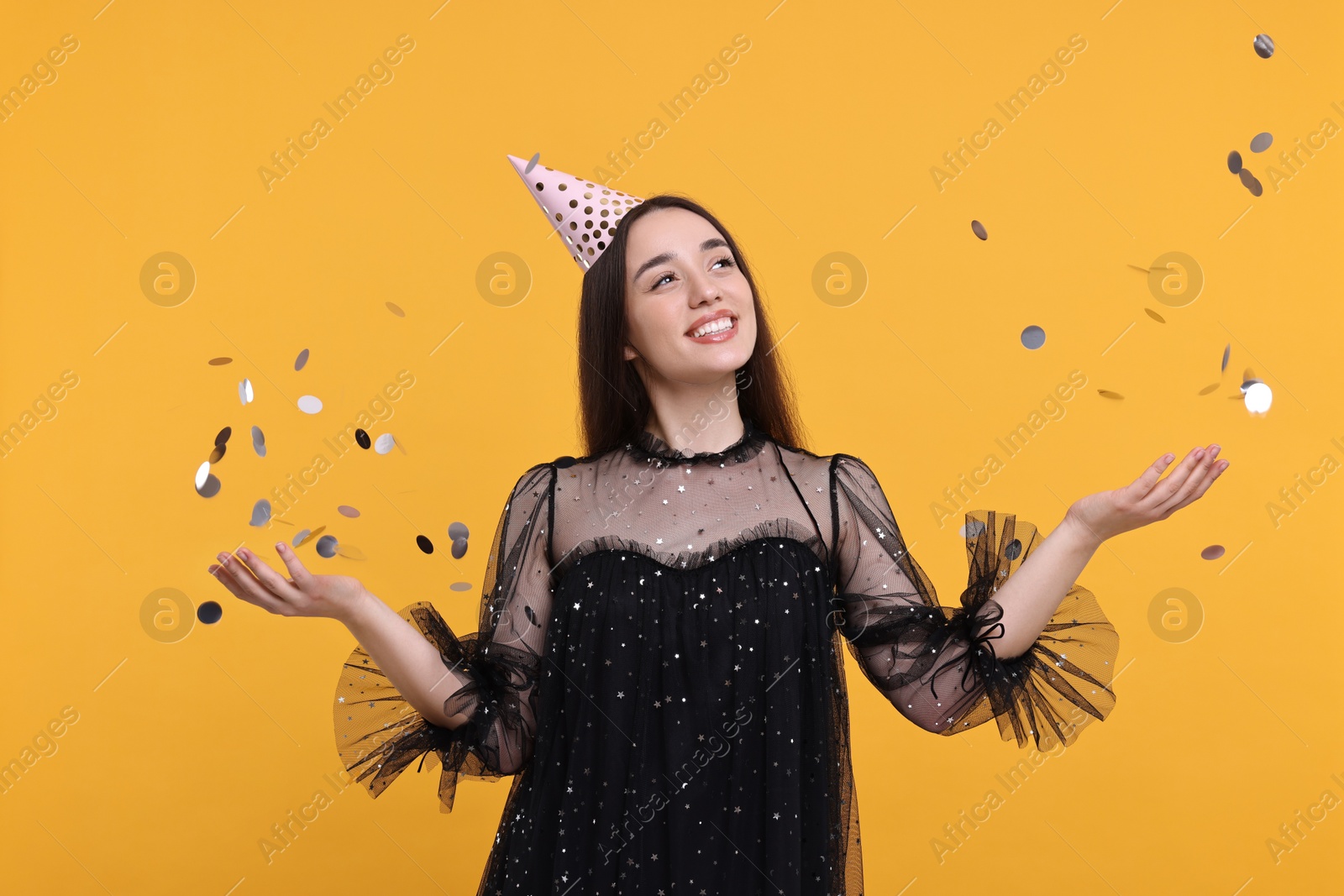 Photo of Happy woman in party hat throwing confetti on orange background