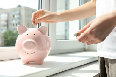 Photo of Woman putting money into piggy bank at window sill indoors, closeup