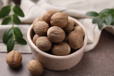 Photo of Whole nutmegs in bowl and green branches on brown table, closeup
