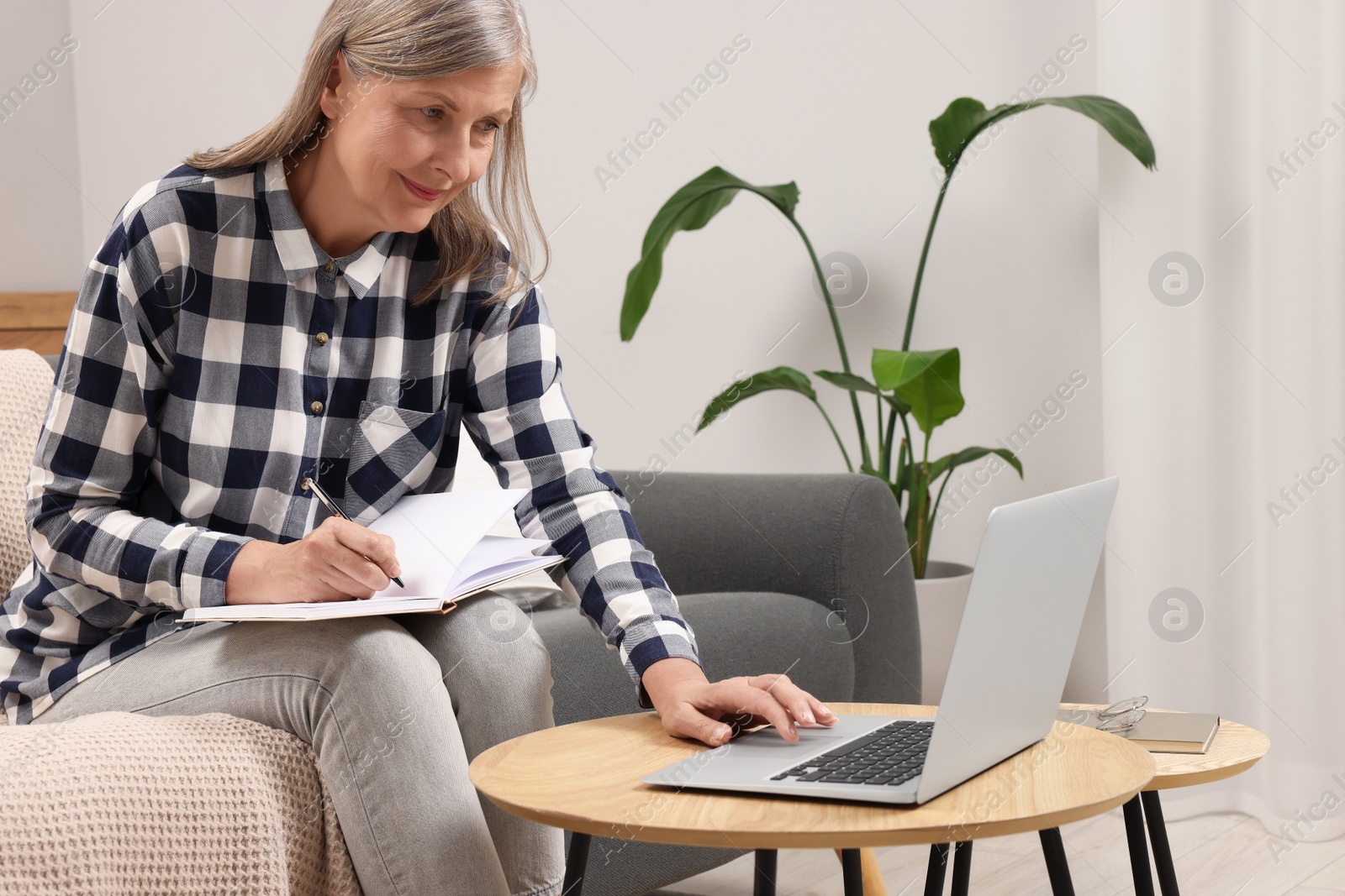 Photo of Beautiful senior woman writing something in notebook while using laptop at home