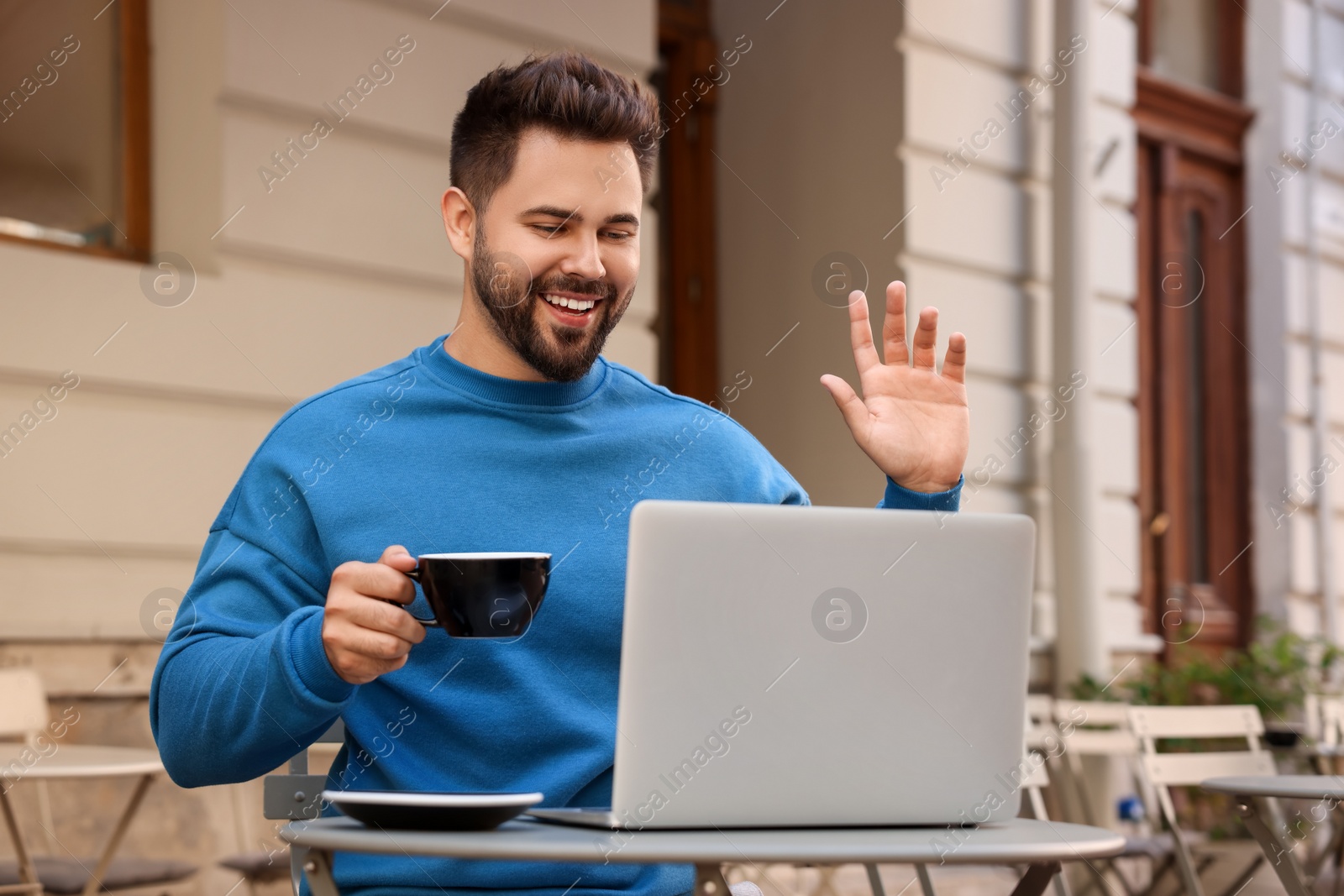 Photo of Handsome young man with cup of drink working on laptop at table in outdoor cafe