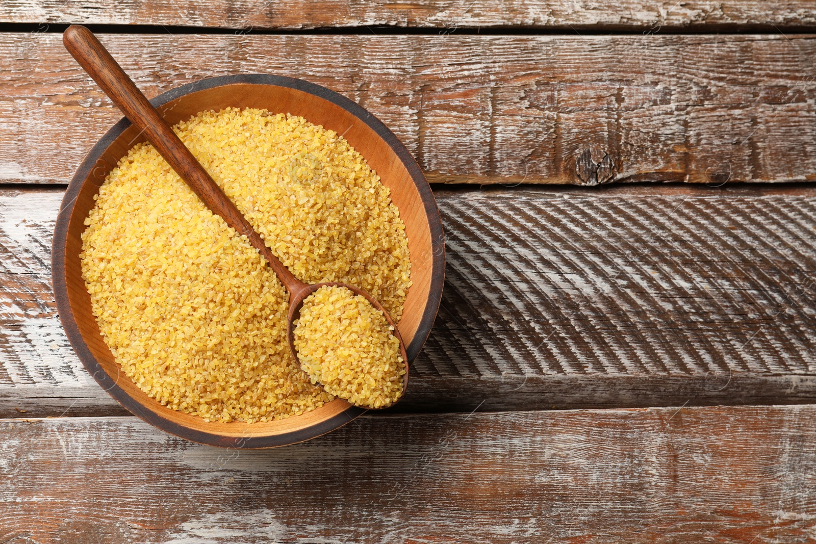 Photo of Bowl and spoon with raw bulgur on wooden table, top view. Space for text