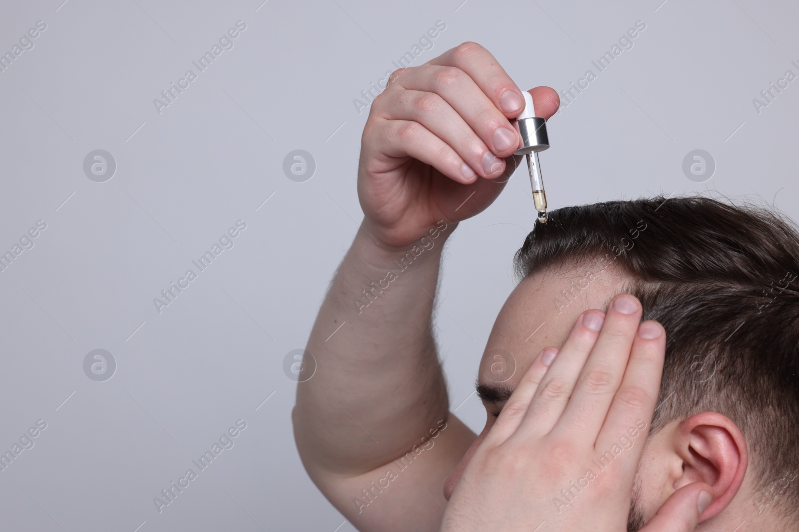 Photo of Baldness concept. Man dripping serum onto his hair on light grey background, closeup. Space for text