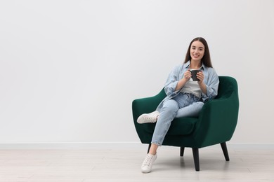 Photo of Beautiful woman with cup of drink sitting in armchair near white wall indoors, space for text