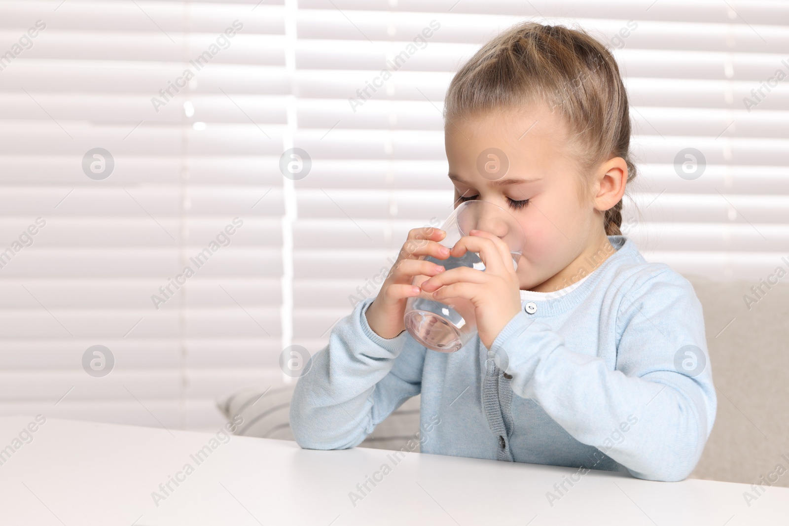 Photo of Cute little girl drinking fresh water from glass at white table indoors
