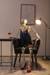 Skeleton in dress with wine sitting at table indoors