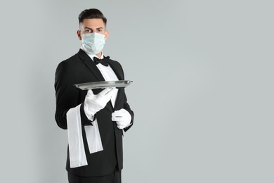 Photo of Waiter in medical face mask with empty tray on light grey background. Space for text