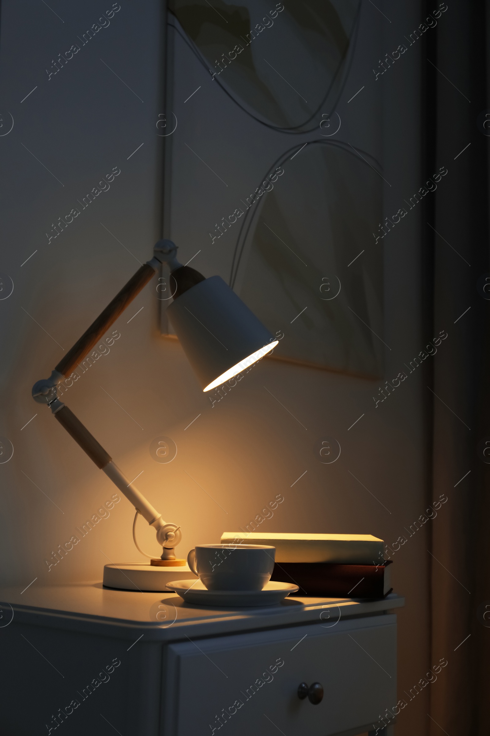 Photo of Stylish lamp, cup of drink and books on table in dark room. Interior design