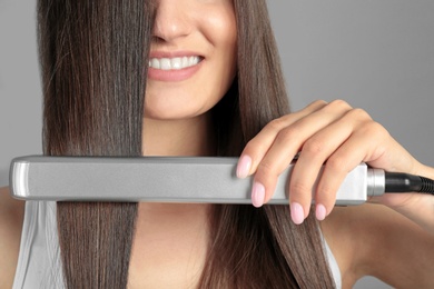 Young woman using hair iron on grey background, closeup