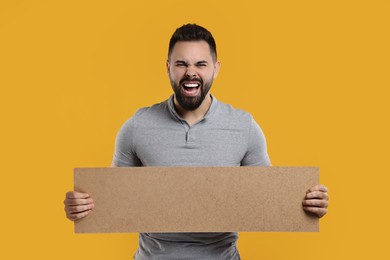 Photo of Angry man holding blank cardboard banner on orange background, space for text