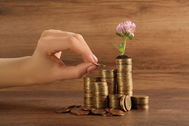 Photo of Woman putting coin onto stack with green plant at wooden table, closeup. Investment concept