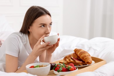 Beautiful woman drinking coffee near tray with breakfast on bed. Space for text