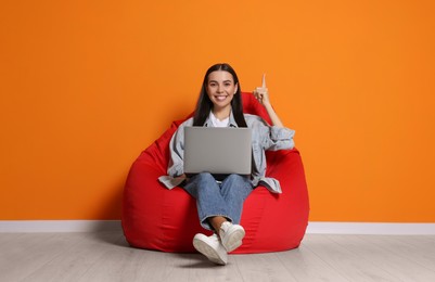 Happy woman with laptop sitting on beanbag chair near orange wall