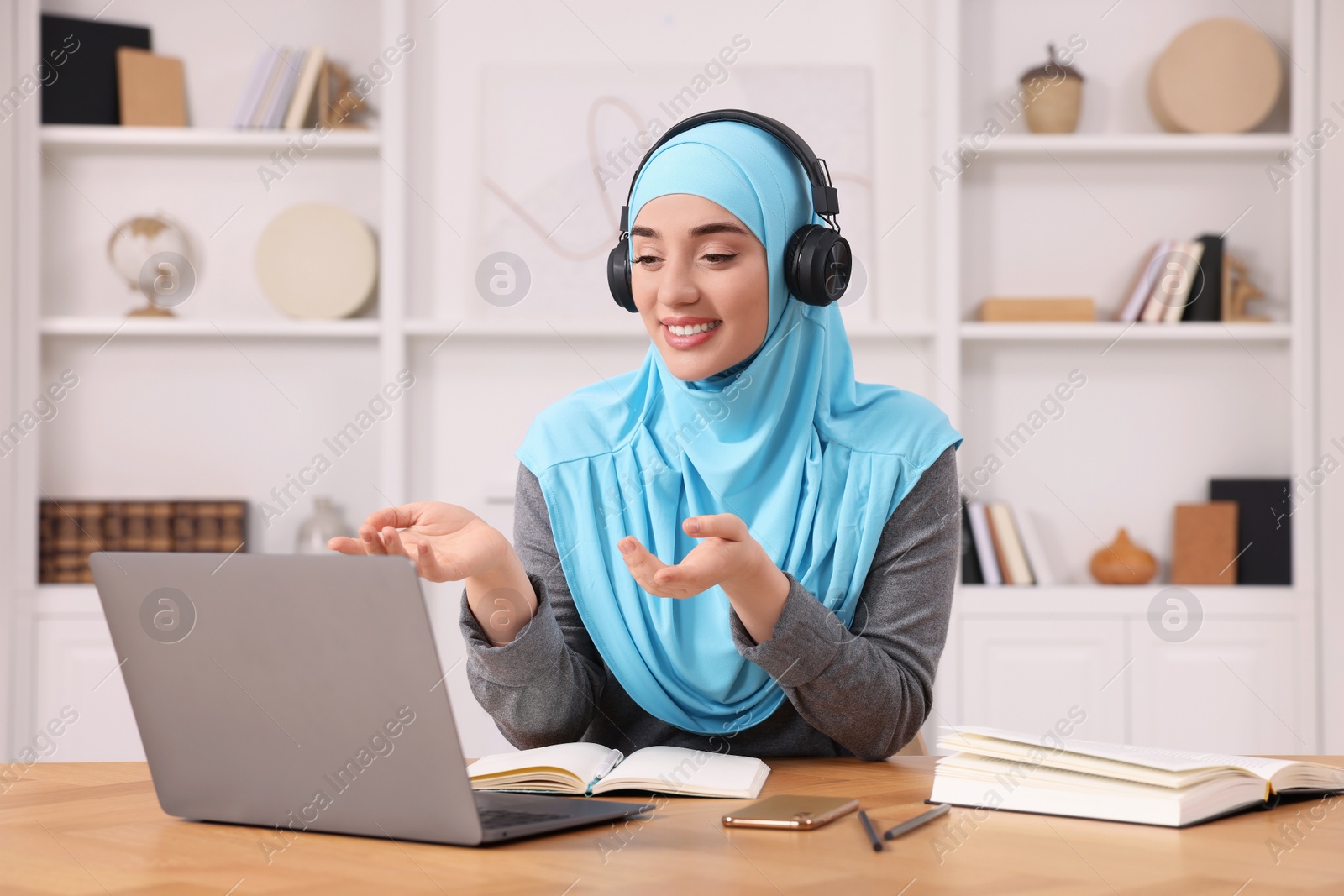 Photo of Muslim woman in hijab using video chat on laptop at wooden table in room