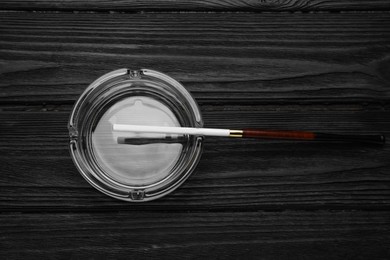 Photo of Cigarette in elegant holder and clean glass ashtray on black wooden table, top view