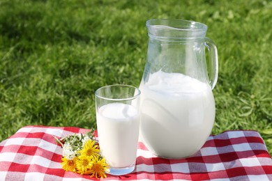 Photo of Jug and glass of fresh milk and flowers on checkered blanket outdoors