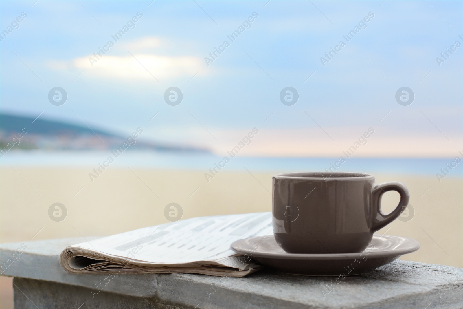 Photo of Ceramic cup of hot drink and newspaper on stone surface near sea in morning. Space for text