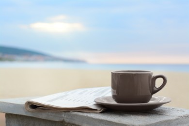Photo of Ceramic cup of hot drink and newspaper on stone surface near sea in morning. Space for text